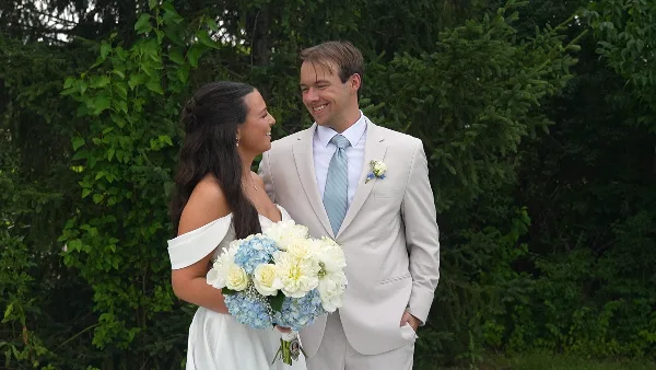 A bride and groom standing outdoors in front of greenery, smiling at each other.