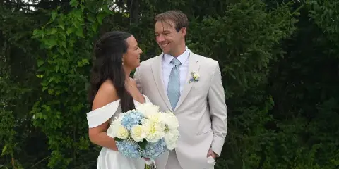 A bride and groom standing outdoors in front of greenery, smiling at each other.