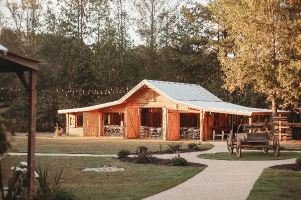 A wide shot of a rustic wooden barn with a metal roof and open doors, set in a grassy field with string lights and a vintage wooden wagon in the foreground.