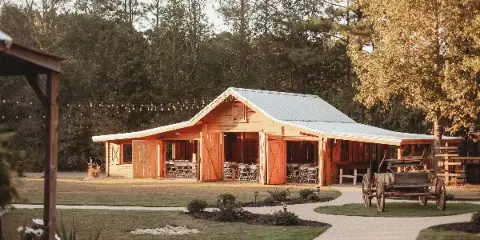 A wide shot of a rustic wooden barn with a metal roof and open doors, set in a grassy field with string lights and a vintage wooden wagon in the foreground.