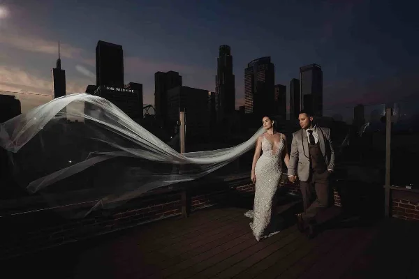 A bride and groom holding hands on a rooftop terrace at dusk with the Los Angeles city skyline in the background.