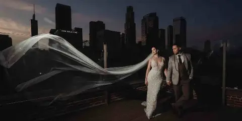 A bride and groom holding hands on a rooftop terrace at dusk with the Los Angeles city skyline in the background.