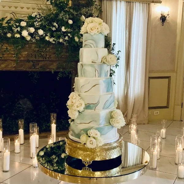 A tall, multi-tiered wedding cake with blue and white marble fondant and white rose decorations, displayed on a gold stand in a venue.