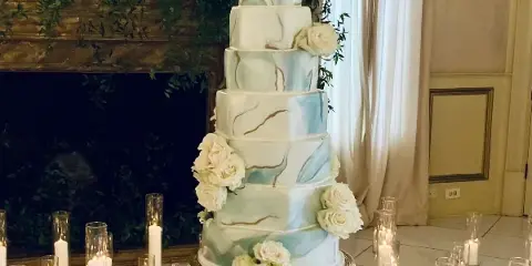 A tall, multi-tiered wedding cake with blue and white marble fondant and white rose decorations, displayed on a gold stand in a venue.