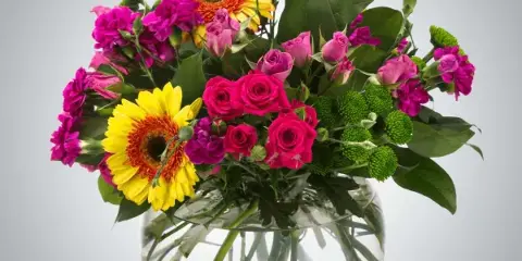A vibrant mixed floral arrangement with pink roses, yellow gerberas, and purple carnations in a clear glass vase.