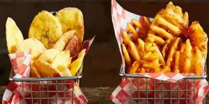 An assortment of prepared food items including sweet potato fries, potato chips, crinkle-cut fries, and green beans served in metal baskets lined with red and white checkered paper.