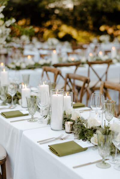 A close-up view of an elegant wedding reception table setting with white linens, tall pillar candles, glassware, and floral centerpieces.
