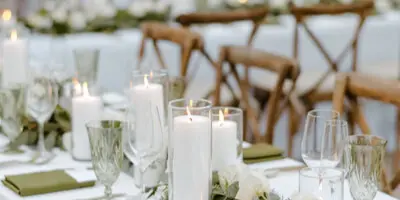 A close-up view of an elegant wedding reception table setting with white linens, tall pillar candles, glassware, and floral centerpieces.