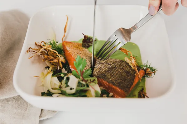 A plated salmon entrée with greens and garnish in a white bowl, being cut with a knife and fork.