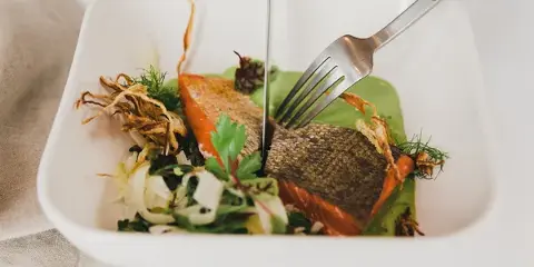 A plated salmon entrée with greens and garnish in a white bowl, being cut with a knife and fork.