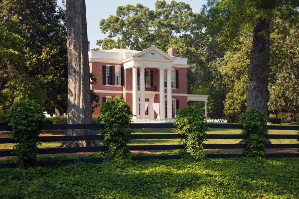 A historic red brick mansion with a white columned portico, set behind a black fence and green lawn.