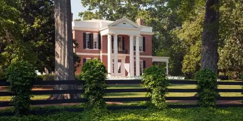 A historic red brick mansion with a white columned portico, set behind a black fence and green lawn.