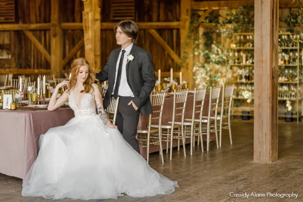 A bride and groom posing inside a rustic wooden barn reception hall with decorated tables and chairs.