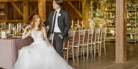 A bride and groom posing inside a rustic wooden barn reception hall with decorated tables and chairs.