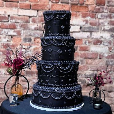 A tall, multi-tiered black wedding cake with intricate silver and white piping, displayed on a table against a brick wall.