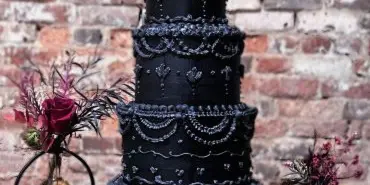 A tall, multi-tiered black wedding cake with intricate silver and white piping, displayed on a table against a brick wall.