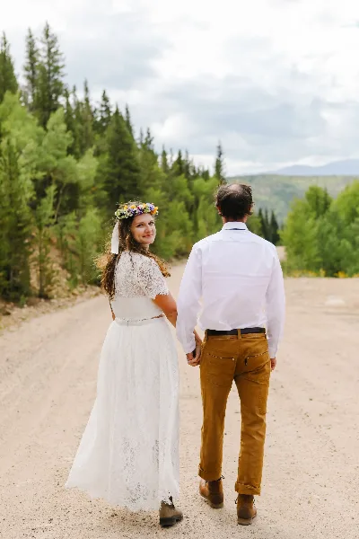 A bride wearing a floral crown and a groom in a white shirt and brown pants walking hand-in-hand on a dirt path surrounded by evergreen trees.