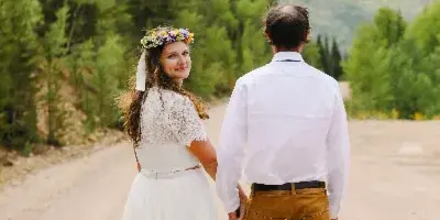 A bride wearing a floral crown and a groom in a white shirt and brown pants walking hand-in-hand on a dirt path surrounded by evergreen trees.