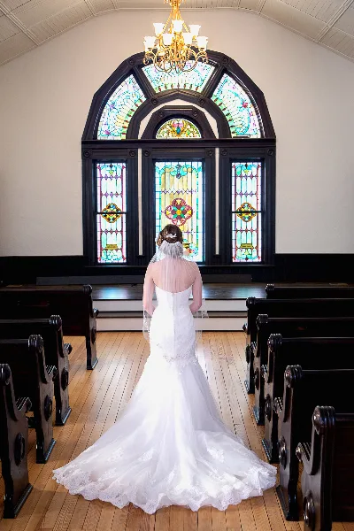 A bride in a white wedding dress standing in the aisle of a chapel with stained glass windows and wooden pews.