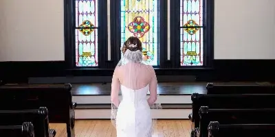A bride in a white wedding dress standing in the aisle of a chapel with stained glass windows and wooden pews.