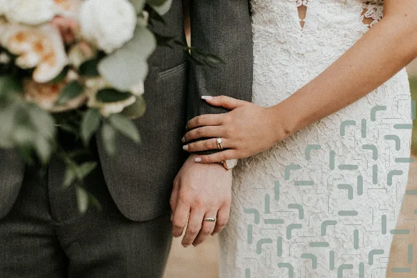 A close-up photograph of a bride and groom standing together, showing the bride's hand with a ring resting on the groom's arm.