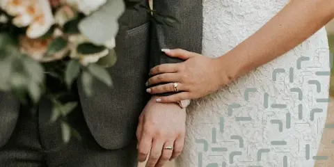 A close-up photograph of a bride and groom standing together, showing the bride's hand with a ring resting on the groom's arm.