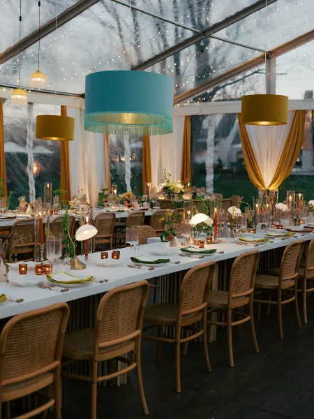A professionally designed wedding reception inside a clear tent featuring a long table, rattan chairs, and colorful hanging pendant lights.
