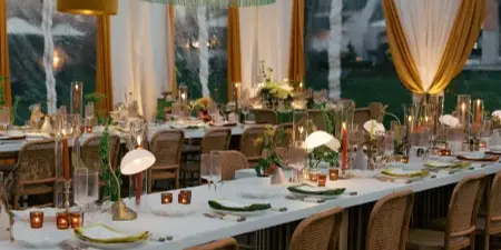 A professionally designed wedding reception inside a clear tent featuring a long table, rattan chairs, and colorful hanging pendant lights.