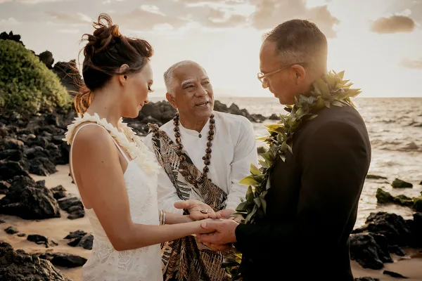 A wedding officiant in traditional attire stands between a bride and groom on a rocky beach, holding their hands during a ceremony.
