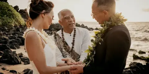 A wedding officiant in traditional attire stands between a bride and groom on a rocky beach, holding their hands during a ceremony.