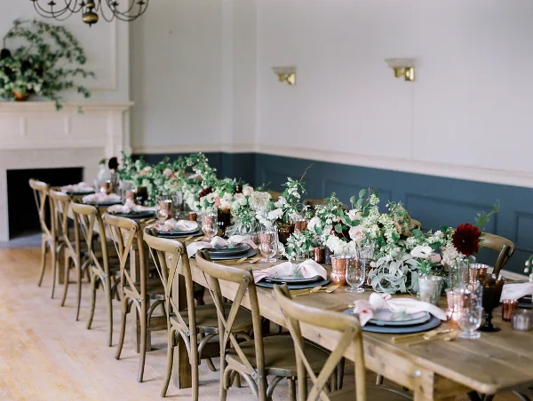 A long wooden dining table set for a wedding with lush floral centerpieces, linens, and curated decor in a rustic venue.