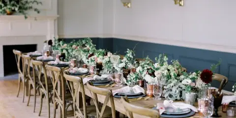 A long wooden dining table set for a wedding with lush floral centerpieces, linens, and curated decor in a rustic venue.
