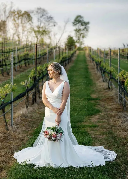 A bride in a white wedding dress and veil stands in a vineyard holding a floral bouquet.