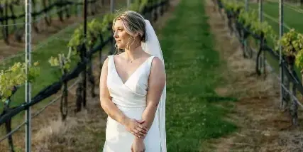A bride in a white wedding dress and veil stands in a vineyard holding a floral bouquet.
