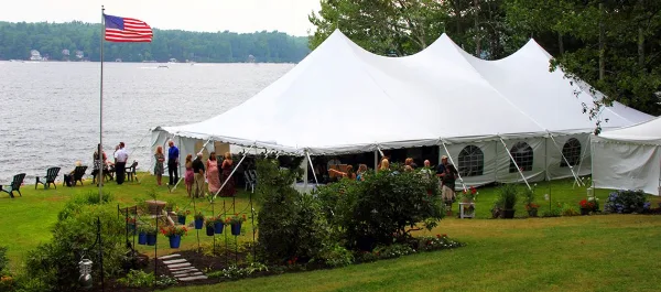 A large white wedding tent set up on a grassy lawn next to a body of water, with guests gathered outside.