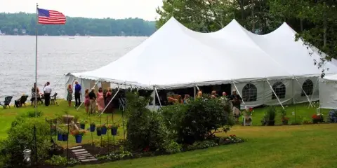 A large white wedding tent set up on a grassy lawn next to a body of water, with guests gathered outside.