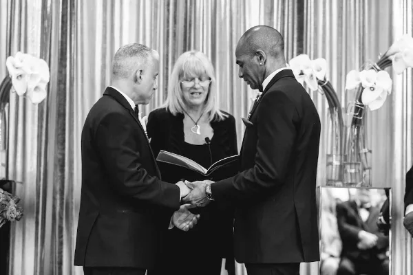 A black and white photograph of a wedding officiant standing between two grooms shaking hands during a ceremony.