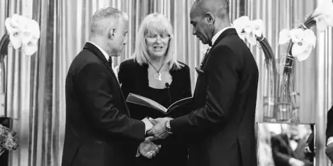 A black and white photograph of a wedding officiant standing between two grooms shaking hands during a ceremony.