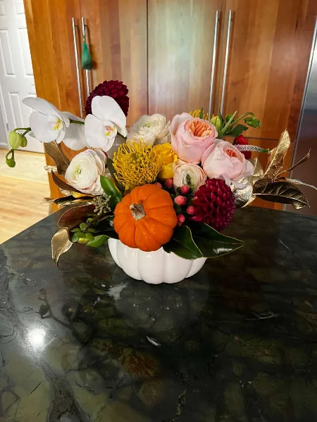 A floral arrangement with white orchids, pink roses, and a small orange pumpkin in a white vase on a dark marble table.