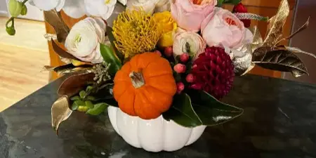 A floral arrangement with white orchids, pink roses, and a small orange pumpkin in a white vase on a dark marble table.