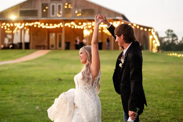 A bride and groom dancing in a grassy field in front of a rustic wooden barn venue decorated with string lights.