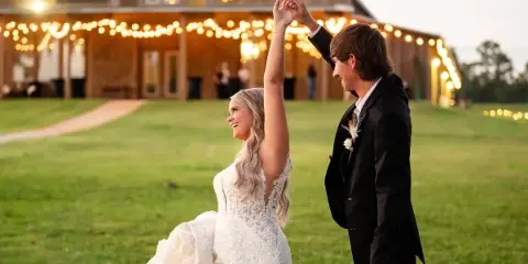 A bride and groom dancing in a grassy field in front of a rustic wooden barn venue decorated with string lights.