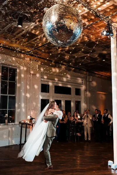 A bride and groom dancing at a wedding reception under a disco ball and string lights, with guests watching and clapping.