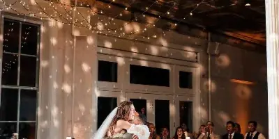 A bride and groom dancing at a wedding reception under a disco ball and string lights, with guests watching and clapping.