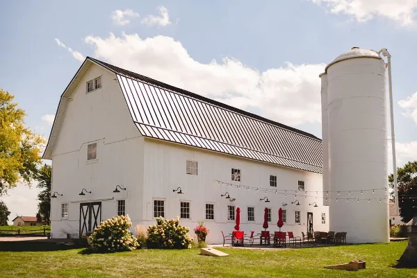 Exterior view of a white barn wedding venue with a silo, outdoor patio seating, and string lights.
