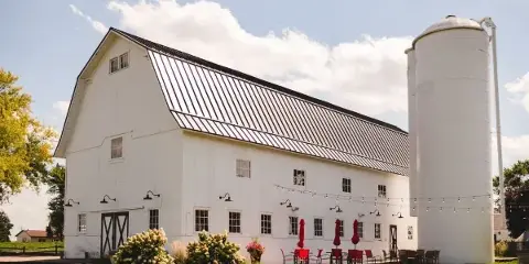 Exterior view of a white barn wedding venue with a silo, outdoor patio seating, and string lights.