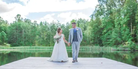 A bride and groom holding hands on a wooden dock extending into a lake, surrounded by a dense forest.