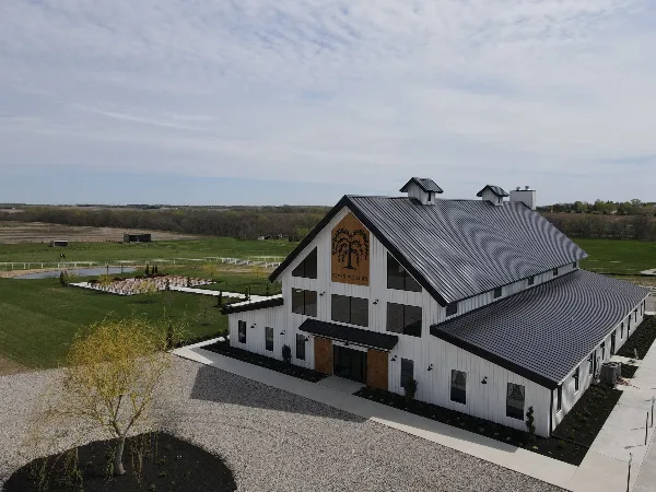 Aerial view of the Seven Willows Venue, a white barn-style building with a dark metal roof, situated on a large green property with a gravel driveway and outdoor seating area.