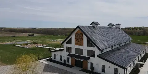 Aerial view of the Seven Willows Venue, a white barn-style building with a dark metal roof, situated on a large green property with a gravel driveway and outdoor seating area.