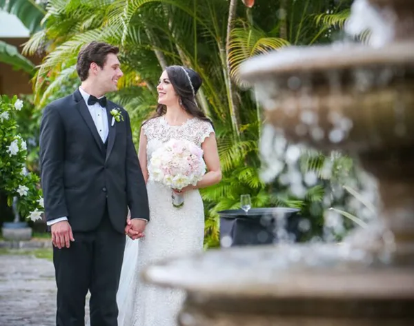 A bride and groom standing together in a tropical garden with a fountain in the foreground.